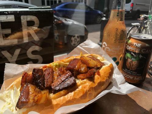 A close-up of a sandwich filled with grilled meat and pineapple, alongside a bottle and can of beverages.