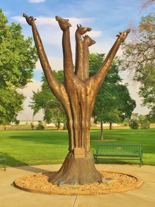 A bronze sculpture of four giraffes with elongated necks, set in a park surrounded by trees and benches.
