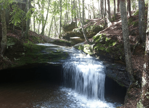 A serene waterfall cascades over moss-covered rocks in a lush, green forest. Sunlight filters through the trees.