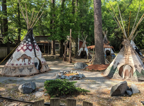A campsite with several colorful tipis surrounded by trees, rocks, and a small fire pit in the center.