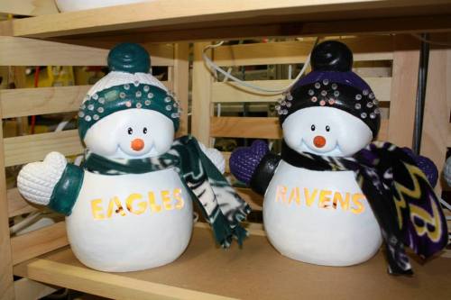 Two snowman figurines wearing scarves and hats, labeled "EAGLES" and "RAVENS," displayed on a wooden shelf.