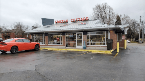 A brick building with a metal roof, featuring a sign that reads "Frozen Custard," and a parked orange car in front.