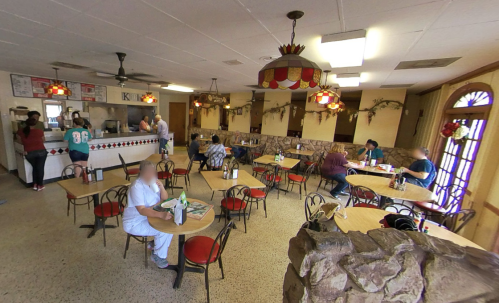 A cozy restaurant interior with tables, chairs, and patrons dining. A counter is visible in the background.