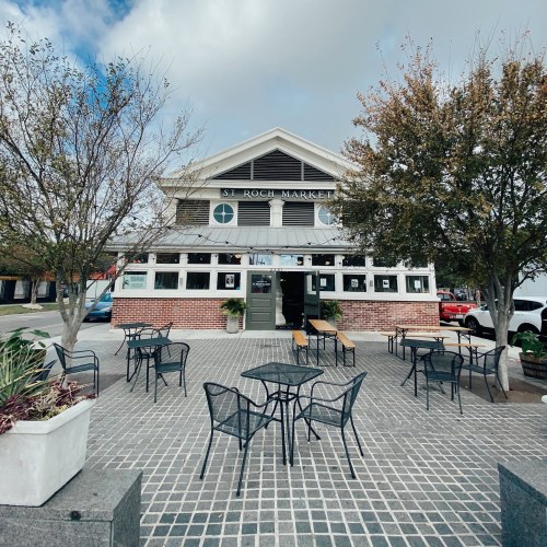 Exterior of St. Roch Market with outdoor seating and trees, under a cloudy sky.