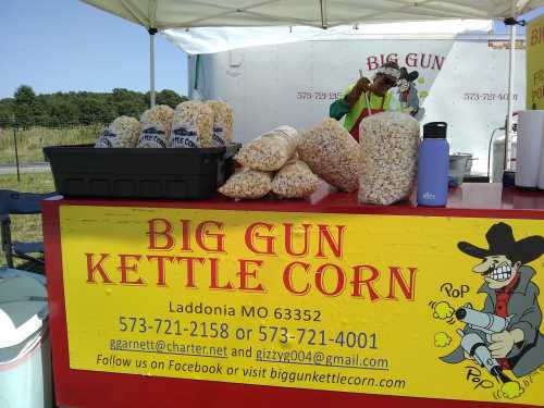 A colorful stand displaying bags of kettle corn, with a sign reading "Big Gun Kettle Corn" and contact information.