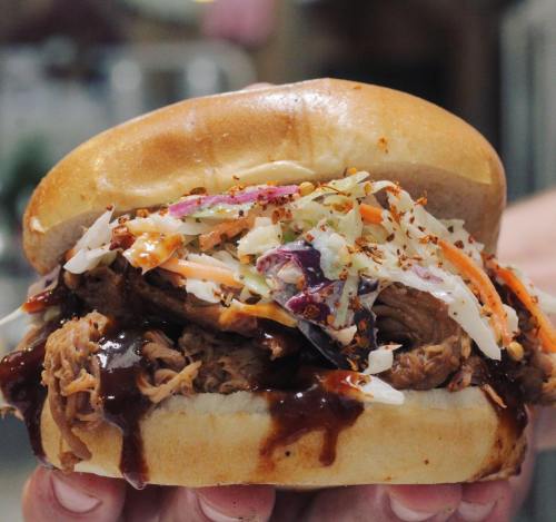 A close-up of a pulled pork sandwich topped with coleslaw and barbecue sauce, held in a hand.