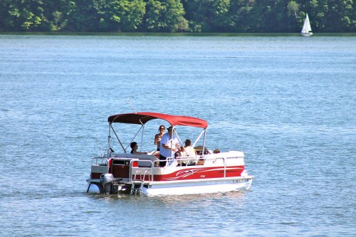 A group of people enjoying a day on a red and white pontoon boat on a calm lake with trees in the background.