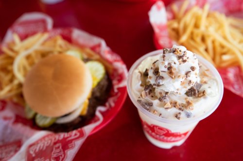 A burger with pickles and a cup of dessert topped with whipped cream and chocolate bits, alongside a serving of fries.