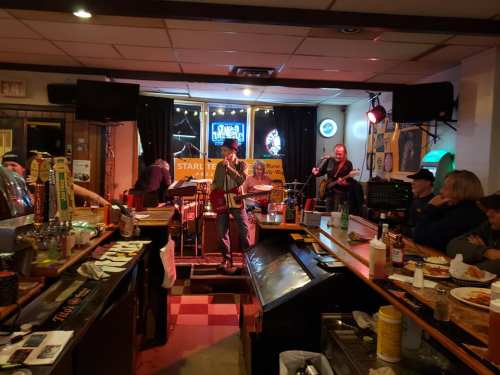 A lively bar scene with a band performing on stage, patrons enjoying drinks and food at the bar.