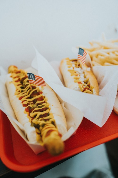 Two hot dogs with mustard, ketchup, and relish, each topped with a small American flag, served on a red tray with fries.