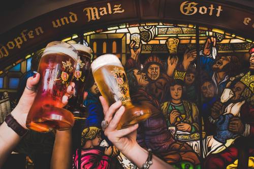 Two hands raise beer glasses in front of a colorful stained glass window depicting a festive gathering.