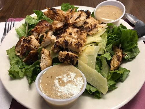 Grilled chicken salad with sliced chicken, lettuce, and two cups of dressing on a white plate.