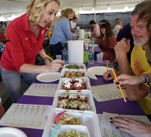 A woman excitedly judges a variety of dishes at a food competition, with participants and tables in the background.