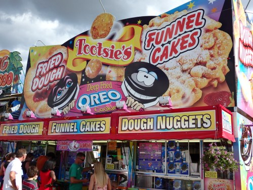 Colorful food stand featuring fried dough, funnel cakes, and deep-fried Oreos at a fair or festival.