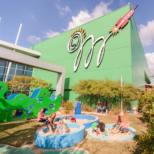 Children playing in sandboxes outside a colorful building with a large mural, surrounded by greenery and blue skies.