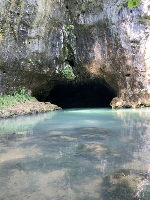 A serene cave entrance by a calm, clear blue water pool surrounded by lush greenery.