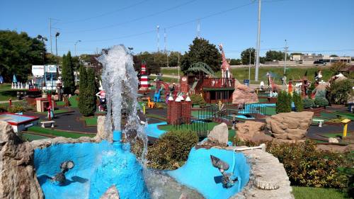 A vibrant mini-golf course featuring colorful obstacles, a fountain, and lush greenery under a clear blue sky.