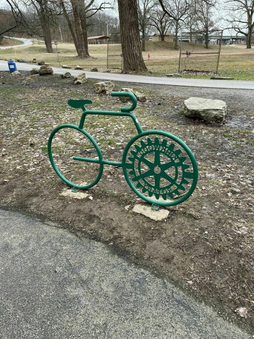 A green bicycle-shaped bike rack made of metal, set on a gravel path in a park.