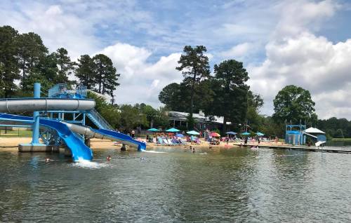 A water park scene with a blue slide, sandy beach, umbrellas, and people enjoying the lake on a sunny day.