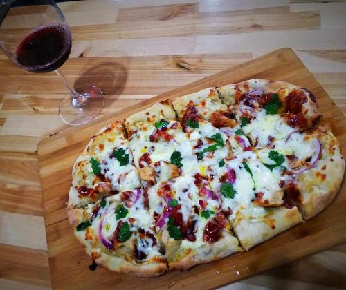 A wooden board with a sliced pizza topped with cheese, chicken, onions, and cilantro, alongside a glass of red wine.