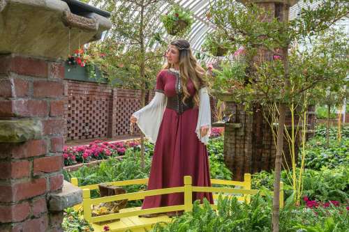 A woman in a flowing red dress stands in a lush garden, surrounded by vibrant flowers and greenery.