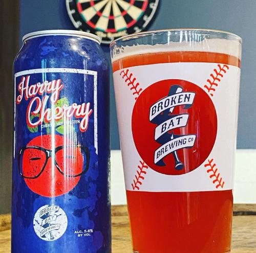 A can of "Harry Cherry" beer next to a glass filled with red liquid, featuring a baseball-themed logo from Broken Bat Brewing.