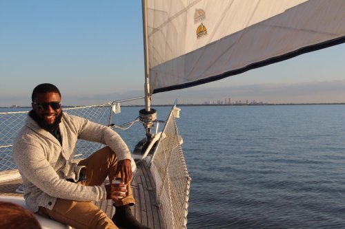 A man in sunglasses relaxes on a sailboat, holding a drink, with a city skyline visible in the background.