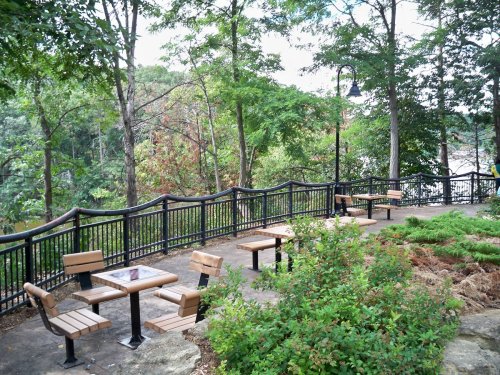 A scenic park area with wooden benches and tables surrounded by trees and a railing overlooking a river.