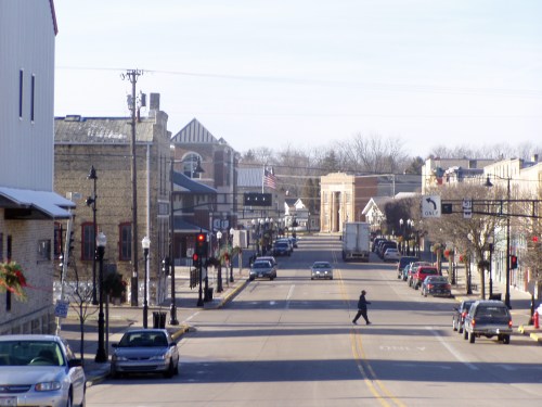 A quiet downtown street with parked cars, a pedestrian, and buildings lining both sides under a clear sky.