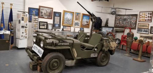 A military jeep displayed in a museum, surrounded by historical artifacts and framed photographs.