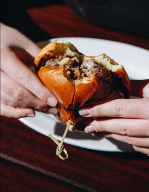 A person holds a half-eaten burger with a golden bun, revealing a juicy patty inside, on a white plate.