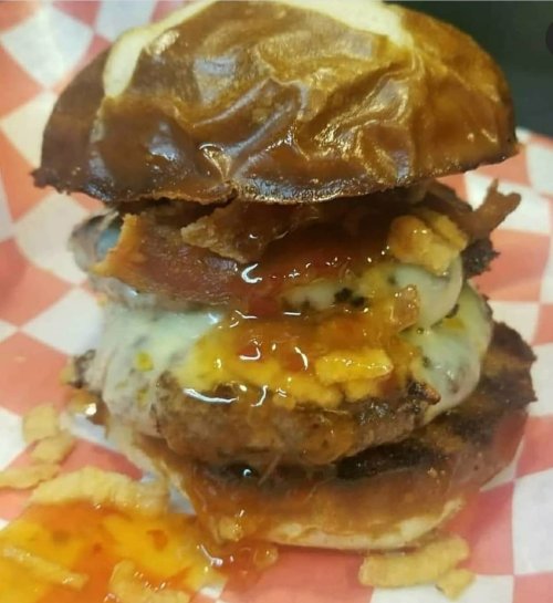 A stacked burger with melted cheese, bacon, and sauce on a pretzel bun, served on a checkered paper.