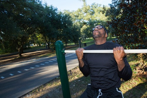 A man performs pull-ups on a bar in a park, surrounded by trees and a quiet road in the background.