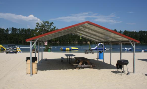A sandy beach area with a covered picnic table, grill, and water activities in the background under a clear blue sky.