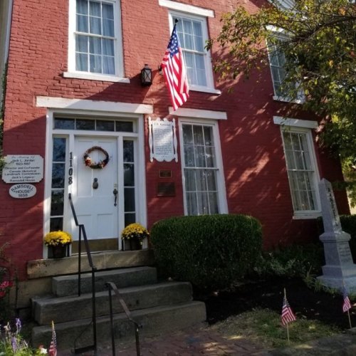 A red brick house with white trim, an American flag, and flower pots on the steps, surrounded by small flags and greenery.