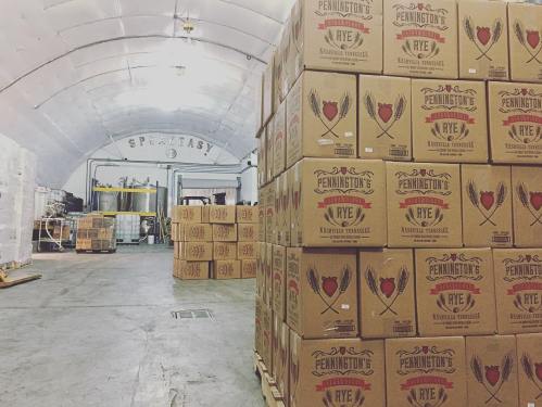 Stacks of Pennington's Rye whiskey boxes in a spacious distillery with a "Speakeasy" sign in the background.