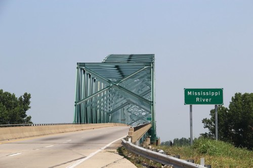 A green metal bridge spans a highway, with a sign indicating the Mississippi River nearby. Clear blue sky above.