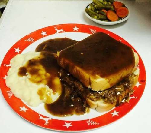 A plate with a thick slice of bread topped with gravy, served with mashed potatoes and a side of green beans and carrots.