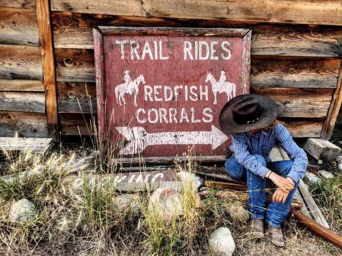 A cowboy in a hat sits beside a weathered sign for trail rides and Redfish Corrals, with tall grass around.