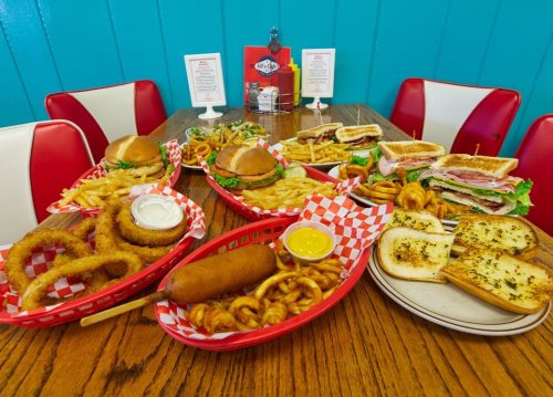 A table filled with burgers, fries, onion rings, a corn dog, pretzels, and sandwiches in a colorful diner setting.