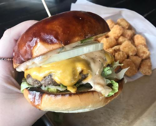 A hand holding a juicy cheeseburger with lettuce, onions, and sauce, alongside a serving of tater tots.
