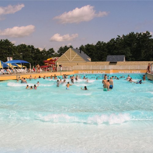 A busy water park with people swimming and playing in a large pool under a blue sky with scattered clouds.
