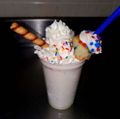 A festive milkshake topped with whipped cream, sprinkles, a mini cupcake, and a striped wafer stick.
