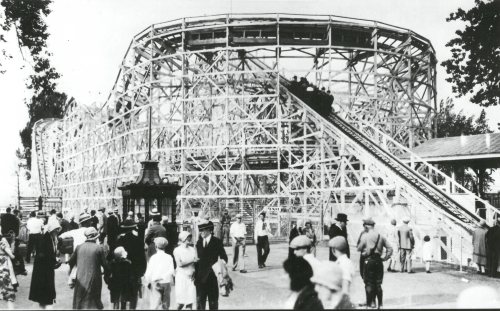 A vintage black-and-white photo of a wooden roller coaster with people walking around in a park setting.