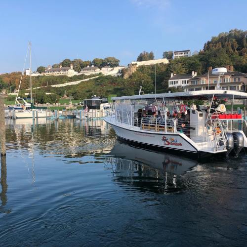 A boat with passengers departs from a dock, surrounded by calm water and scenic hills with houses in the background.