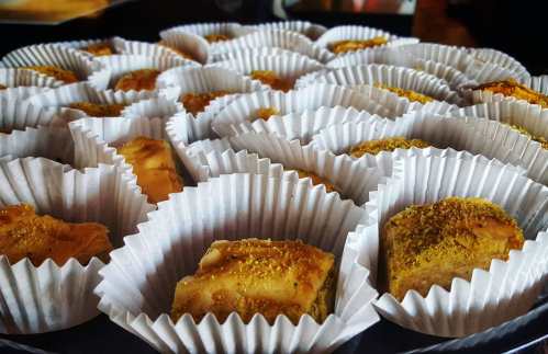 A close-up of assorted baklava pieces arranged in white paper cups, topped with a sprinkle of golden pistachio.