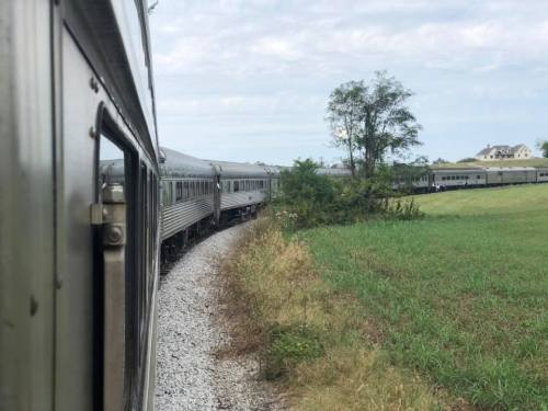 A train curves around a bend, passing through a grassy landscape under a cloudy sky.