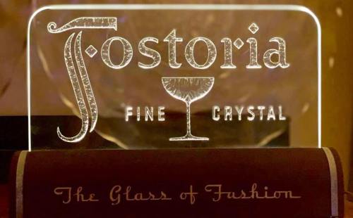 Illuminated sign for Fostoria Fine Crystal, featuring a glass icon and the tagline "The Glass of Fashion."