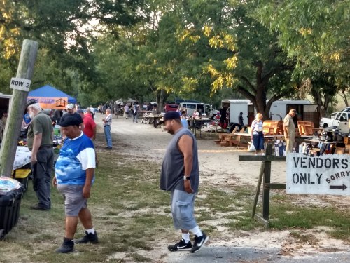 A bustling outdoor market scene with vendors and visitors walking along a dirt path lined with trees and tents.