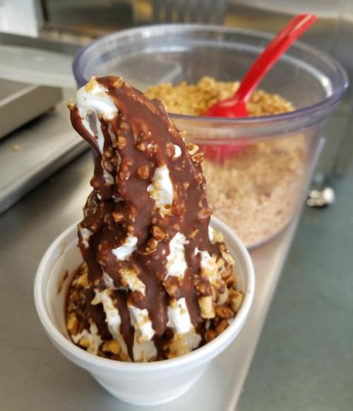 A soft-serve ice cream cone topped with chocolate and crunchy toppings, served in a cup with a bowl of granola in the background.